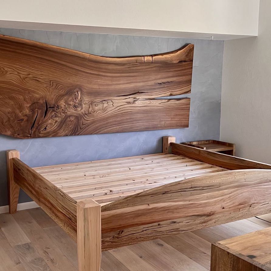 Live edge slab headboard on a blue plaster wall above a solid wood bed frame and matching nightstands