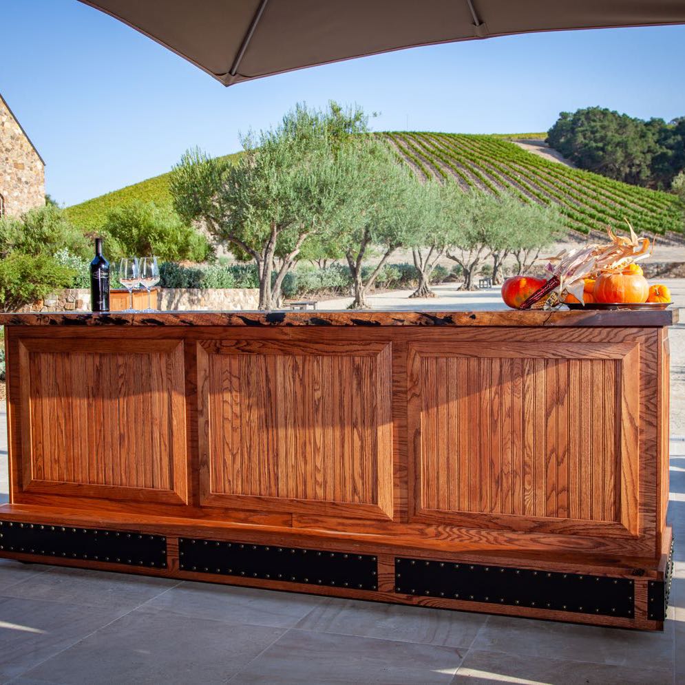 Paneled wood bar with a live edge top on a winery patio, with a vineyard hill behind