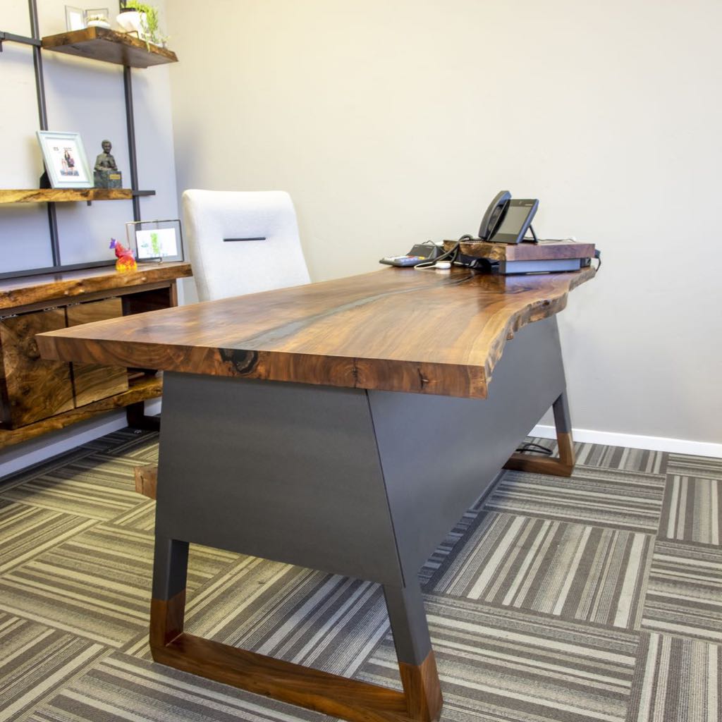 Live edge desk on a dark steel base with wood feet in an office, with live edge shelves behind
