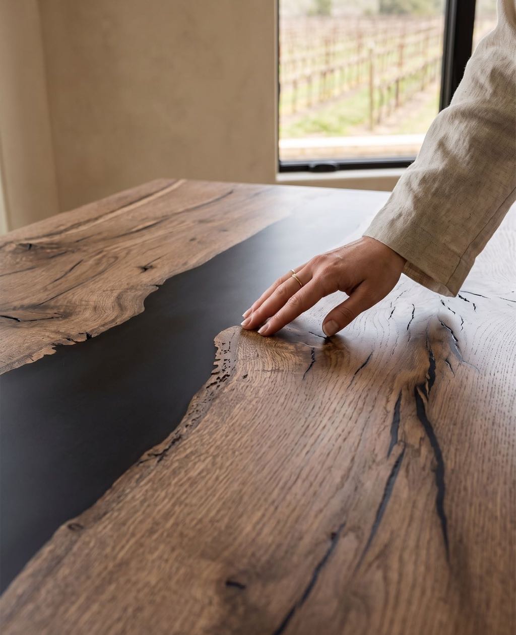A hand resting on the live edge of a coast live oak river table, with vineyard rows through the window