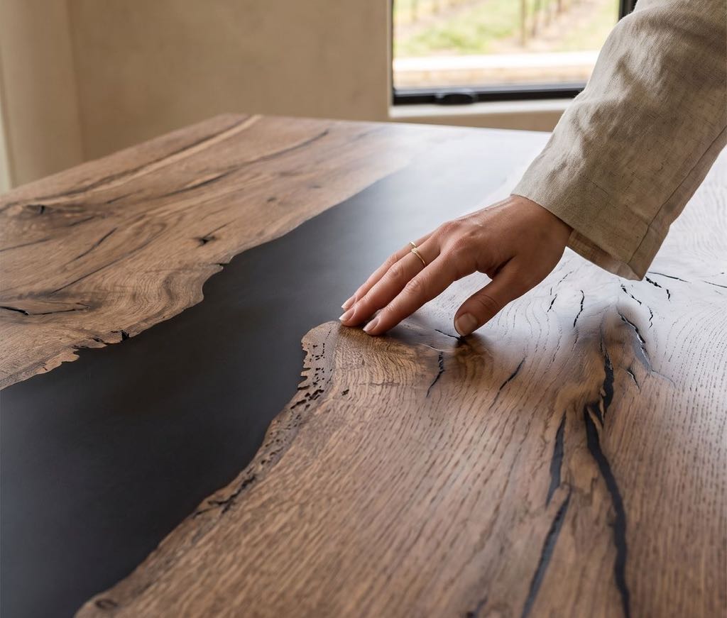 A hand resting on the oak top beside the black river, with vineyard rows through the window