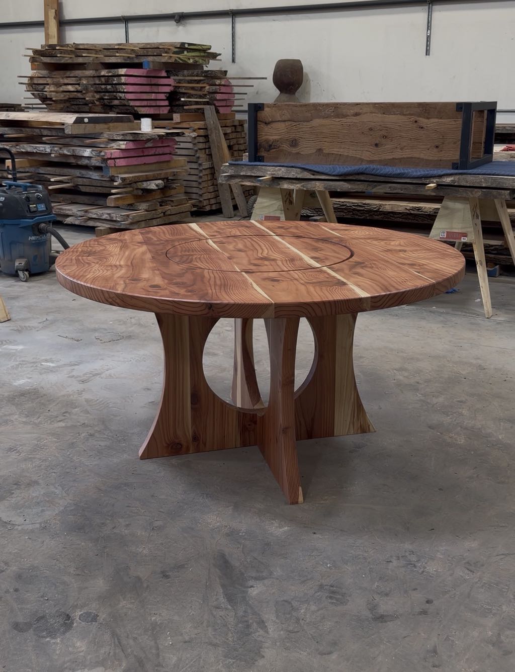 The finished round redwood table standing on the shop floor, with lumber racks behind it