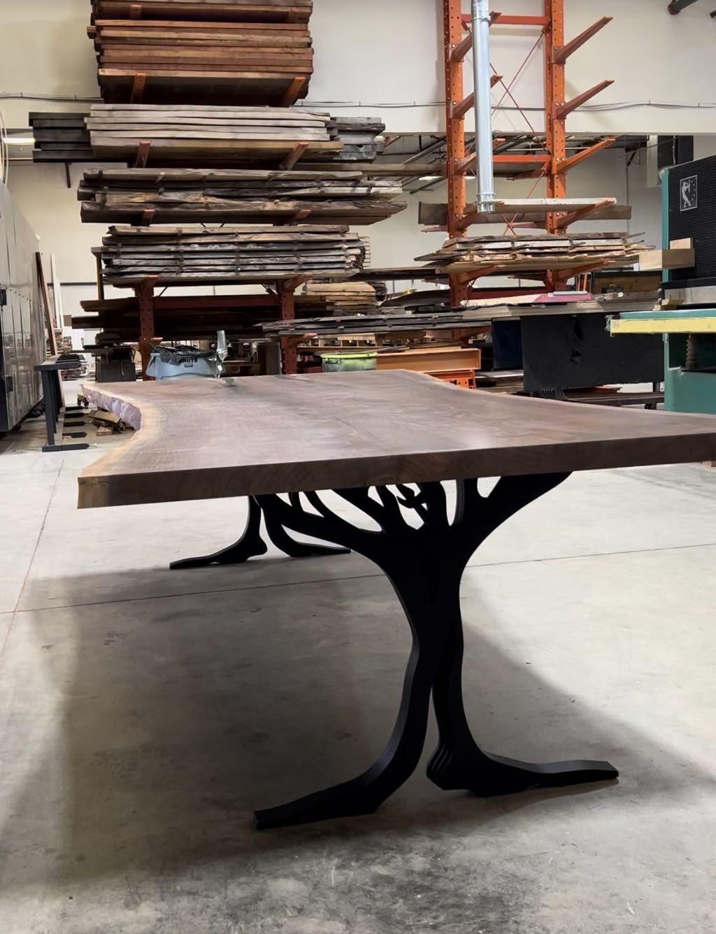 The finished table on the shop floor, seen low from one end, with both tree legs in view and lumber racks behind