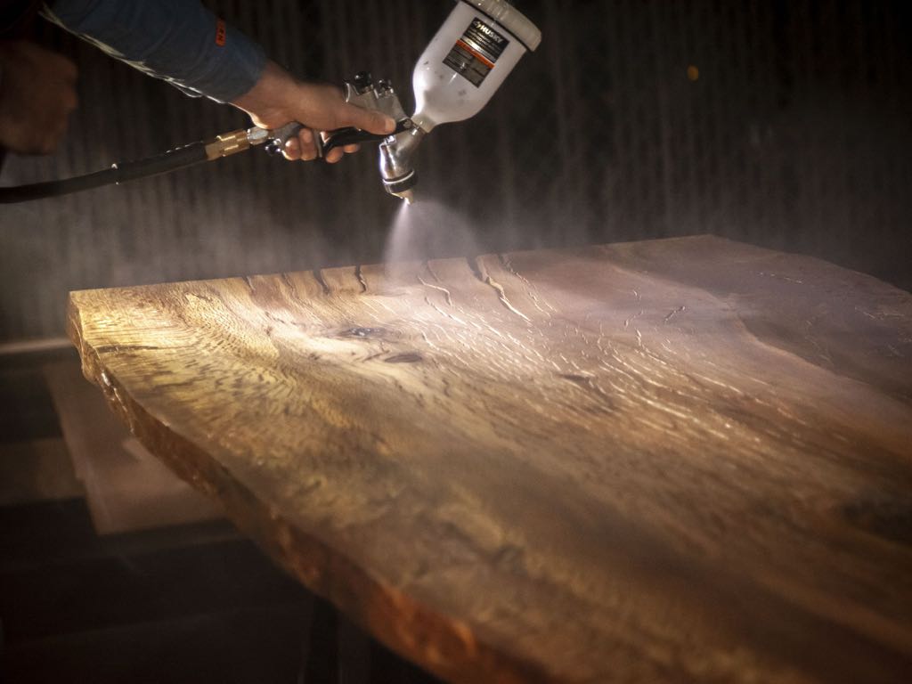 A spray gun laying finish across a wide slab, lit against a dark shop