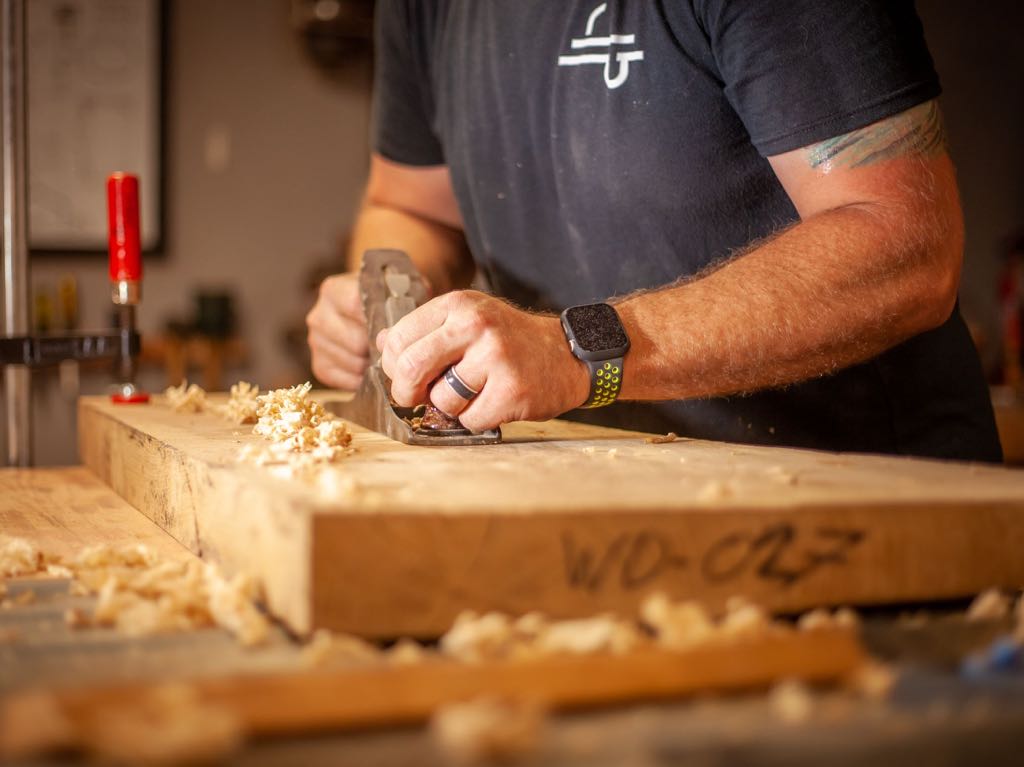 Hands pushing a hand plane along the edge of a thick board, with curls of shavings on the bench