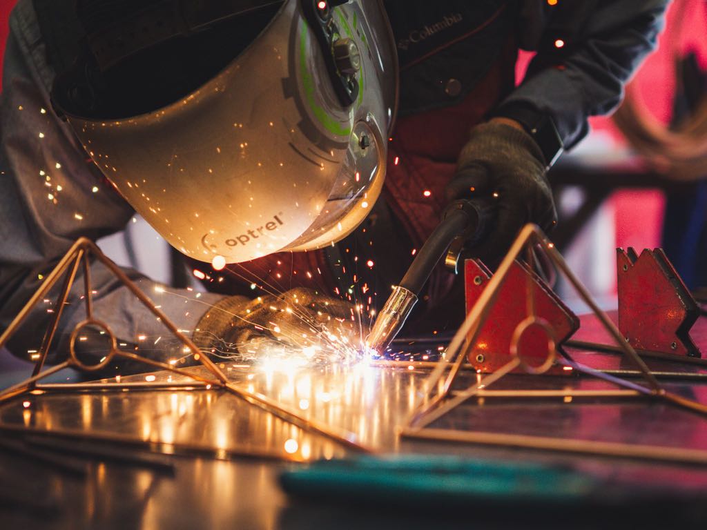 A welder in a helmet joining a steel frame, with sparks flying off the bench