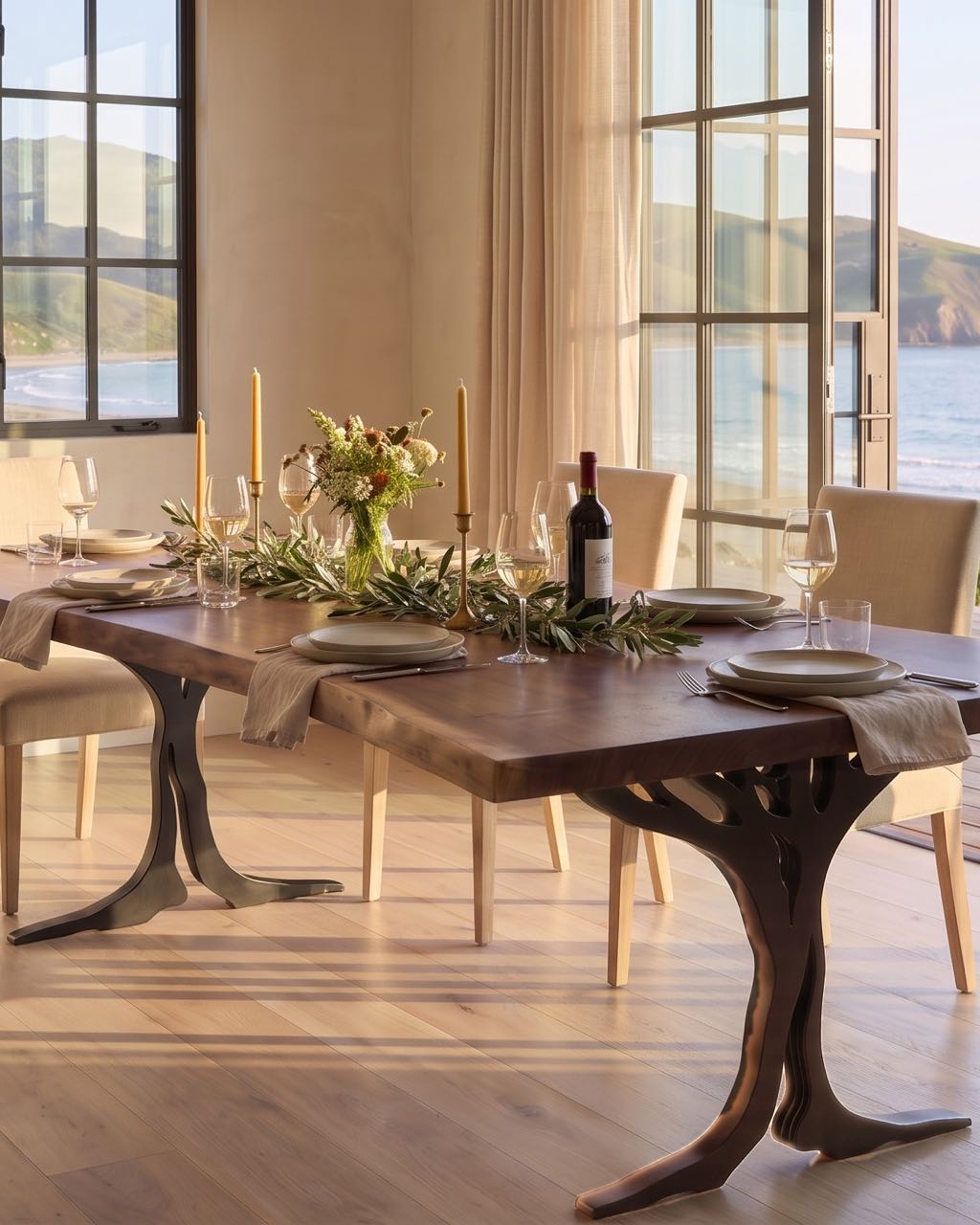 Claro walnut dining table on two black steel tree-shaped legs, set for dinner in a styled room with ocean views at golden hour