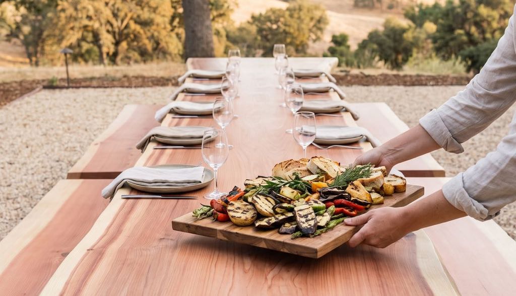 Hands setting a board of grilled vegetables on the redwood table at an outdoor dinner under an oak