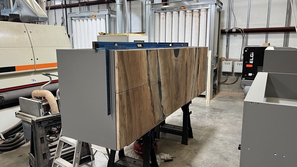 The credenza fronts being fitted to the gray carcass in our shop, with black resin running through the seams between the sycamore panels and a level on top